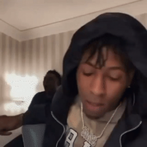 A Man Wearing A Hoodie And A Necklace Is Sitting In A Room With His Eyes Closed . GIF