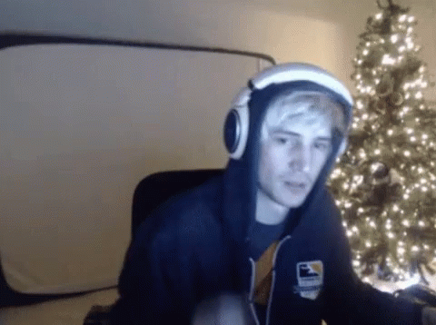 A Man Wearing A Hoodie And Headphones Is Sitting In Front Of A Christmas Tree .. GIF