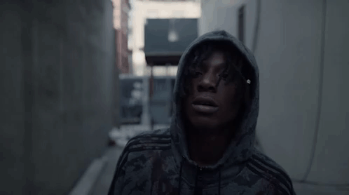 A Man Wearing A Hoodie Is Standing In A Dark Alleyway . GIF