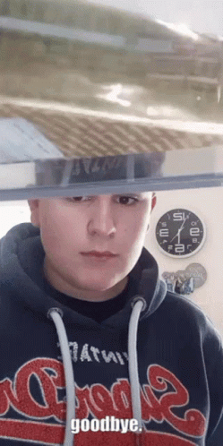 A Man Wearing A Hoodie That Says Goodbye Stands In Front Of A Clock GIF