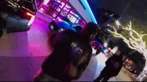 A Man Wearing A Hoodie With A Skull And Crossbones On The Back Walks Down A Street At Night GIF