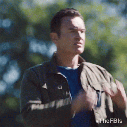 a man wearing a jacket and a blue shirt is standing in the woods and making a funny face .