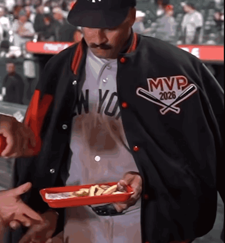A Man Wearing A Jacket That Says Mvp 2026 Holds A Tray Of Food GIF