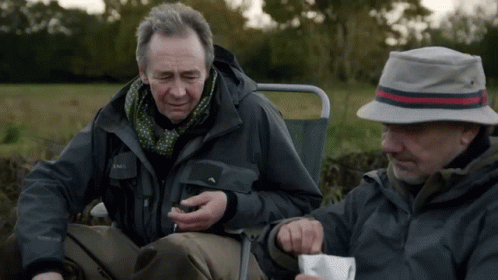A Man Wearing A Jacket That Says ' Salomon ' On It Sits Next To Another Man Wearing A Hat GIF