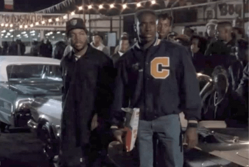 A Man Wearing A Jacket With The Letter C On It Stands Next To Another Man GIF