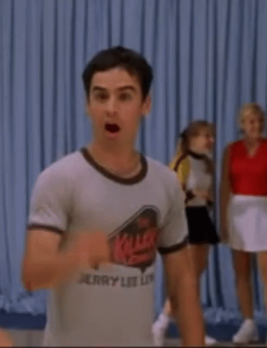 A Man Wearing A Jerry Lee Lip T-Shirt Is Standing In Front Of A Blue Curtain . GIF