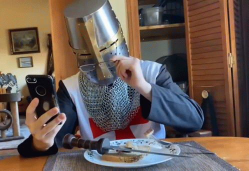 A Man Wearing A Knight 'S Helmet Is Sitting At A Table Eating Bread And Looking At His Phone GIF