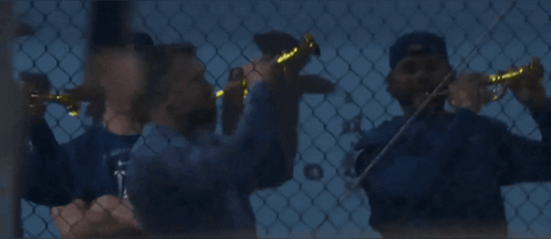 A Man Wearing A La Hat Stands Behind A Chain Link Fence While Another Man Plays A Violin GIF