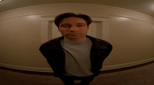 A Man Wearing A Leather Jacket And A Grey Shirt Stands In A Hallway GIF