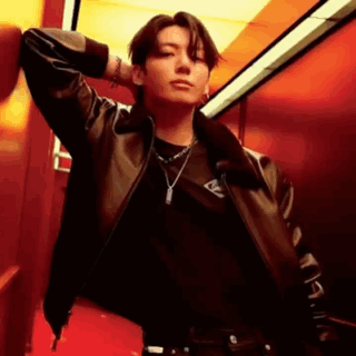 A Man Wearing A Leather Jacket And A Necklace Is Leaning Against A Red Wall . GIF