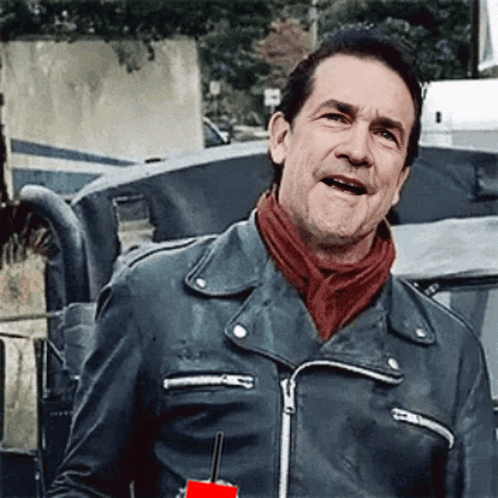 A Man Wearing A Leather Jacket And A Red Scarf Is Standing In Front Of A Vehicle . GIF