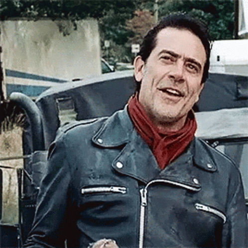 A Man Wearing A Leather Jacket And A Scarf Is Standing In Front Of A Car . GIF
