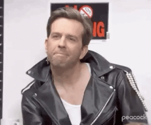 A Man Wearing A Leather Jacket And A White Shirt Is Sitting In Front Of A No Smoking Sign . GIF