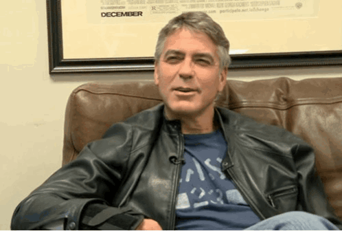A Man Wearing A Leather Jacket Sits On A Couch In Front Of A Framed Picture That Says December GIF