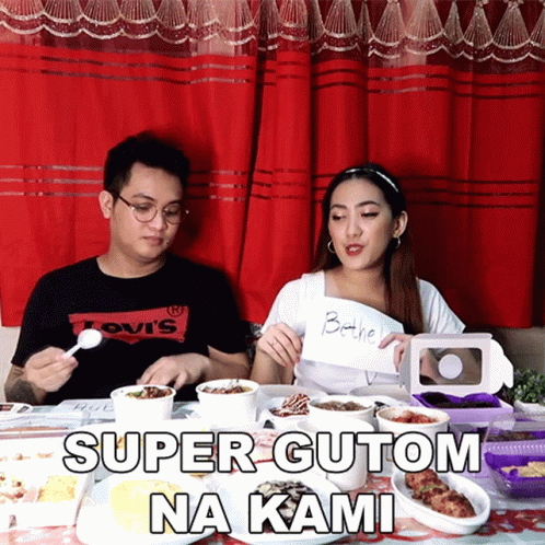 A Man Wearing A Levis Shirt Sits At A Table With A Woman Holding A Sign That Says Super Gutom Na Kami GIF