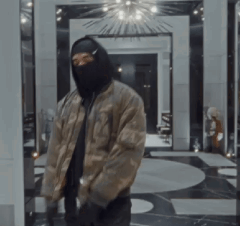 A Man Wearing A Mask And A Bomber Jacket Is Standing In A Room . GIF