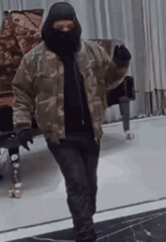 A Man Wearing A Mask And A Camouflage Jacket Is Standing In A Room . GIF