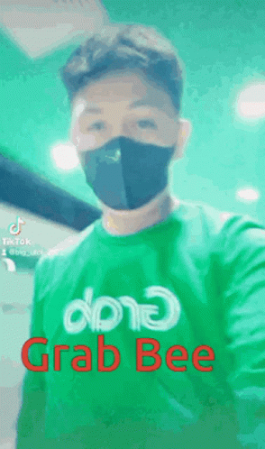 A Man Wearing A Mask And A Green Shirt That Says Grab Bee Is Taking A Selfie . GIF
