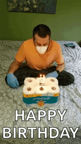 A Man Wearing A Mask And Gloves Is Sitting On A Bed With A Birthday Cake Made Out Of Toilet Paper Rolls . GIF