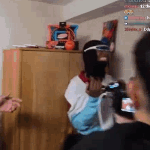 A Man Wearing A Mask And Goggles Is Standing In A Room Holding A Remote Control . GIF