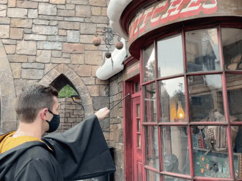 A Man Wearing A Mask Holds A Wand In Front Of A Witches Shop GIF