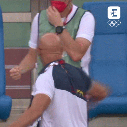A Man Wearing A Mask Is Being Helped By Another Man With The Olympic Logo In The Background GIF