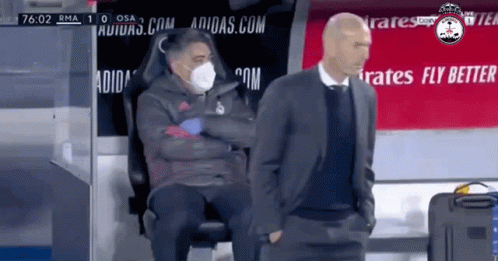 A Man Wearing A Mask Is Sitting Next To Another Man In A Suit GIF