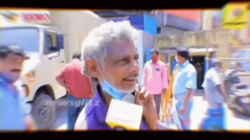 A Man Wearing A Mask Is Talking Into A Microphone While Standing In A Crowd . GIF