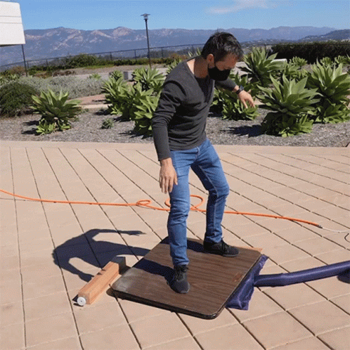 A Man Wearing A Mask Stands On A Wooden Table Outside GIF