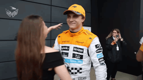 A Man Wearing A Mclaren Racing Suit Talks To A Woman GIF