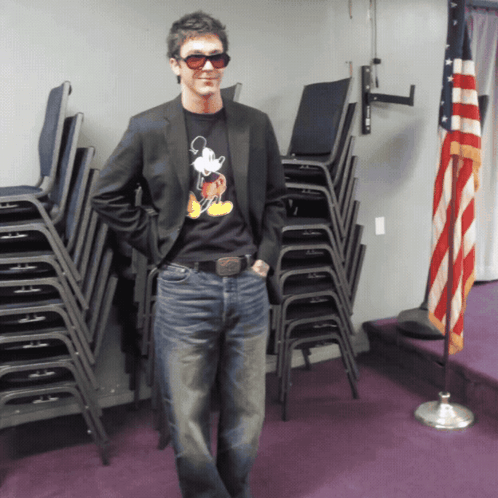 a man wearing a mickey mouse shirt stands in front of stacks of chairs