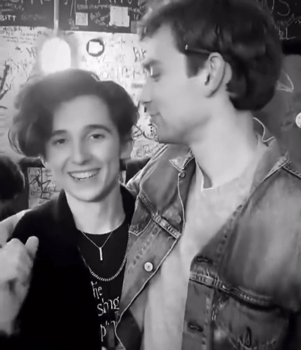 A Man Wearing A Necklace That Says ' The Singer ' On It Stands Next To Another Man GIF