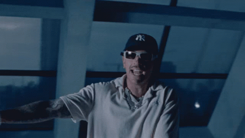 A Man Wearing A Ny Hat And Sunglasses Stands In Front Of A Window GIF