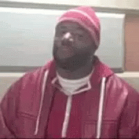 A Man Wearing A Pink Hat And A Red Jacket Is Sitting In A Chair . GIF