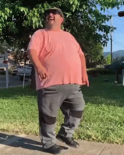 A Man Wearing A Pink Shirt And Grey Pants Is Standing On A Sidewalk GIF