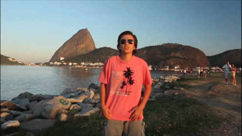 A Man Wearing A Pink Shirt With Palm Trees On It Stands In Front Of A Body Of Water GIF