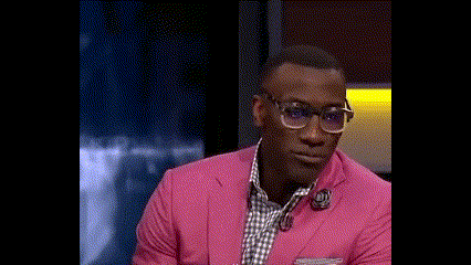 A Man Wearing A Pink Suit And Glasses Is Smiling While Sitting On A Stage . GIF