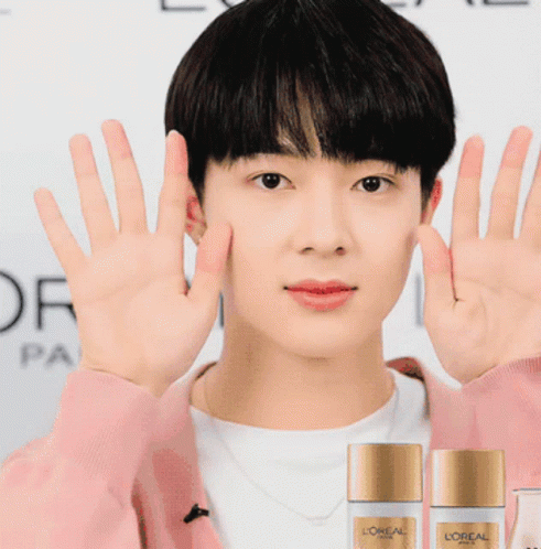 A Man Wearing A Pink Sweater Holds Up His Hands In Front Of Two Bottles Of L' Oreal Paris GIF