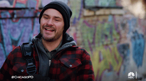 A Man Wearing A Plaid Jacket And A Beanie Is Smiling In Front Of A Graffiti Covered Wall .. GIF