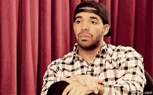 A Man Wearing A Plaid Shirt And A Baseball Cap Is Sitting In Front Of A Red Curtain GIF