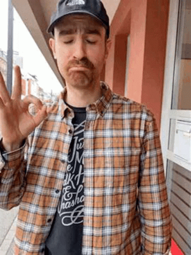 A Man Wearing A Plaid Shirt And A Black Hat Is Giving The Ok Sign . GIF