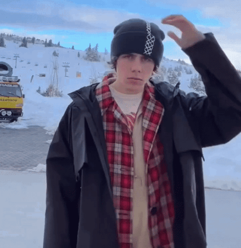 A Man Wearing A Plaid Shirt And A Black Jacket Stands In Front Of A Snowy Mountain GIF