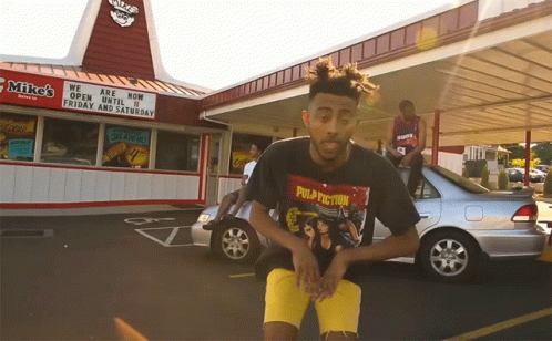 A Man Wearing A Pulp Fiction T-Shirt Stands In Front Of A Mike 'S Restaurant GIF