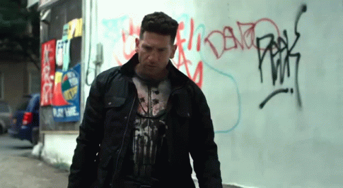 A Man Wearing A Punisher T-Shirt Stands In Front Of A Graffiti Covered Wall GIF