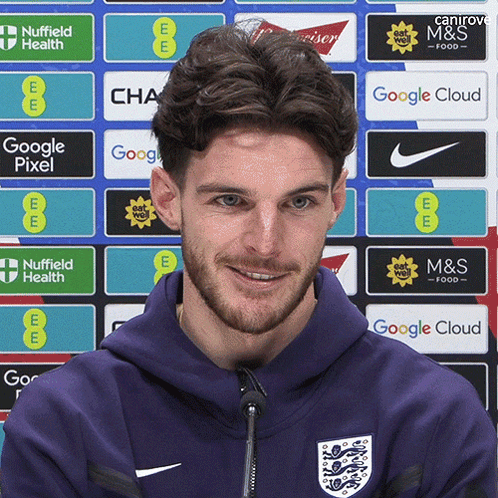 A Man Wearing A Purple Hoodie Stands In Front Of A Wall Of Advertisements Including Google Pixel And M & S Food GIF