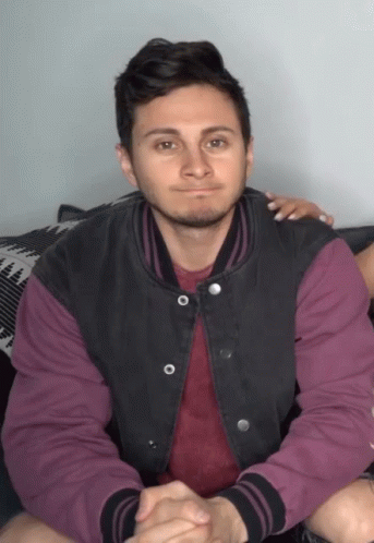 A Man Wearing A Purple Shirt And A Black Jacket Is Sitting On A Couch GIF