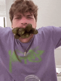 A Man Wearing A Purple Shirt With A Green Letter K On It Is Eating Pickles GIF