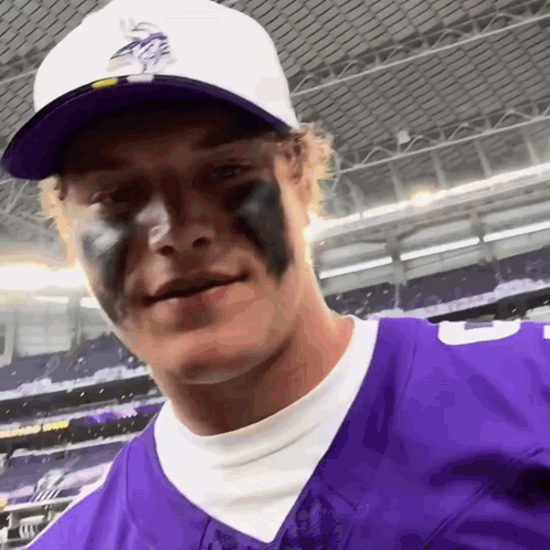 A Man Wearing A Purple Vikings Jersey And A White Hat Has Black Paint On His Face GIF