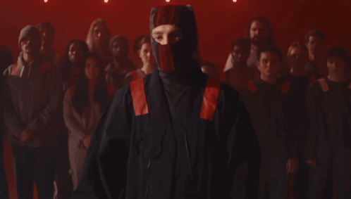A Man Wearing A Red And Black Mask Stands In Front Of A Crowd Of People GIF