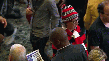 A Man Wearing A Red And Black Striped Hat Is Reading A Newspaper In A Crowd Of People GIF